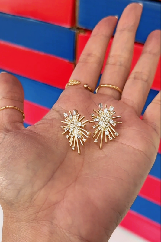 A close-up of a person's hand holding a pair of gold starburst stud earrings with clear rhinestones, with gold rings on the fingers, against a red and blue striped background.
