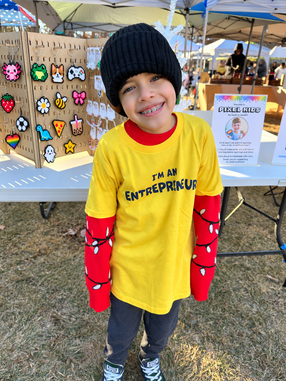 A young child smiling at an outdoor market, wearing a black knit beanie, a bright yellow long-sleeve shirt with 'I'M AN ENTREPRENEUR' printed across the chest, and red long-sleeve sleeves decorated with black string lights.