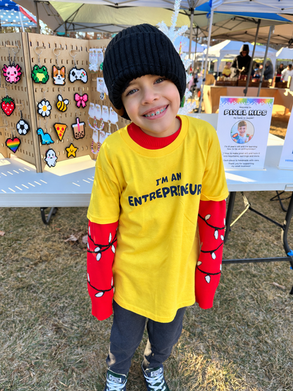 A young child smiling at an outdoor market, wearing a black knit beanie, a bright yellow long-sleeve shirt with 'I'M AN ENTREPRENEUR' printed across the chest, and red long-sleeve sleeves decorated with black string lights.